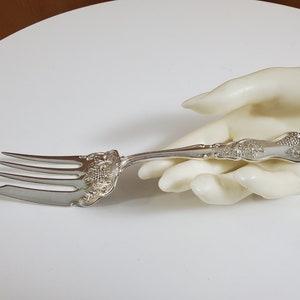 Peut inclure: Une fourchette de service en argent avec un motif orné de raisins et de feuilles. La fourchette a trois dents et un long manche.