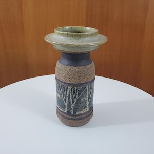 Peut inclure: Un vase en céramique avec un large dessus plat et un corps cylindrique. Le dessus est de couleur vert clair, la section du milieu est bleu foncé et le corps est décoré d'un motif d'arbres dans des tons de vert et de marron. Le vase est sur une table blanche.