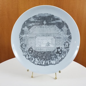 May include: A white ceramic plate featuring a black and white illustration of a building with a stage and a crowd of people in front of it. The illustration is surrounded by a black border.