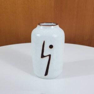 May include: A small white ceramic vase with a brown abstract design. The vase is approximately 10 cm tall and has a slightly rounded shape.