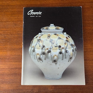 May include: A ceramic jar with a lid is featured on the cover of "Ceramics Monthly" magazine. The jar is white with a speckled pattern of brown and yellow. The magazine cover is set against a dark background.
