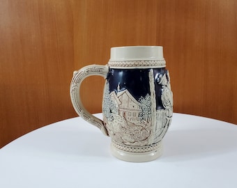 May include: A ceramic beer stein with a blue and white design featuring a small house and a figure in traditional clothing. The stein has a handle and a raised rim.