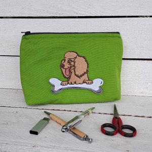 May include: Green zippered pouch with a brown and white embroidered dog sitting on a bone. The dog has a pink tongue sticking out.