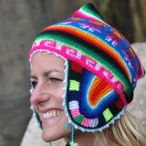 Può includere: Una donna indossa un cappello di lana colorato con le alette per le orecchie e i pon pon. Il cappello presenta un motivo arcobaleno ed è realizzato con una varietà di colori, tra cui rosa, blu, verde, giallo e rosso.