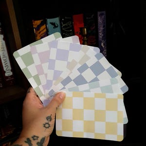 May include: A hand holding a fan of rectangular cards with a checkered pattern. The cards display various pastel colors, including yellow, green, blue, and purple. The background is a dark bookshelf with colorful book spines.
