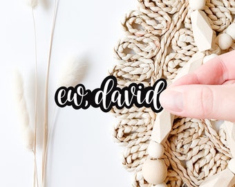 May include: A black vinyl sticker with the text "ew, david" in a cursive font. The sticker is being held by a hand over a woven straw mat with wooden beads.