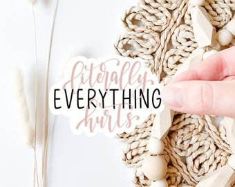 May include: White sticker with black and pink lettering that reads "literally EVERYTHING hurts". The sticker is being placed on a woven straw mat with wooden beads.