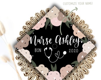 May include: Black graduation cap with a floral design in pink and beige. The text "Nurse Ashley BSN 2020" is written on the cap with a stethoscope and heart graphic.