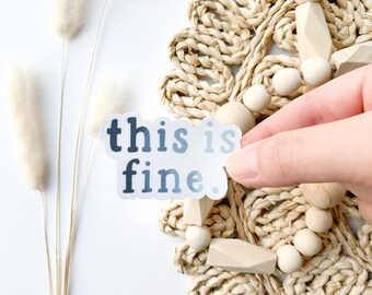 May include: A white sticker with the text "this is fine." in black text with a blue gradient. The sticker is being held by a hand over a woven straw mat with wooden beads.