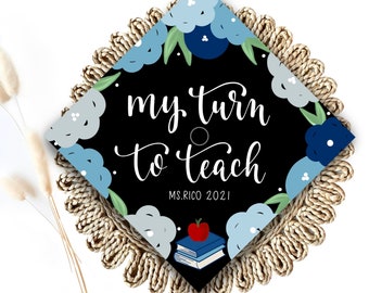 May include: A black graduation cap with a blue and white floral design. The text "my turn to teach" is written in white cursive lettering. The text "MS. RICO 2021" is written in white lettering below the cursive text. A stack of books with a red apple on top is pictured below the text.