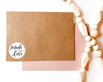 May include: A brown paper envelope with a white circle sticker that says "made with love" in black script with pink hearts. The envelope is on a white surface with a wooden bead garland.