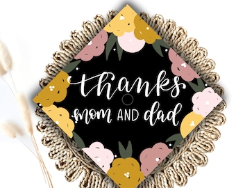 May include: A black diamond-shaped card with a floral design in pink, yellow, and green. The card says "Thanks Mom and Dad" in white cursive lettering.