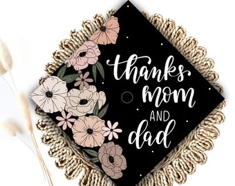 May include: Black graduation cap with a floral design and the text "thanks mom and dad" in white cursive lettering.