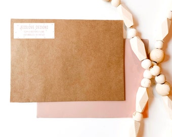 May include: A brown paper envelope with a white return address label that reads "Jesslove Designs 1234 E Butterfly Lane Los Angeles, CA 90745". The envelope is on a white surface with a white wooden bead garland.