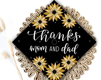 May include: Black graduation cap with gold sunflowers and the text "Thanks mom and dad" in white cursive lettering.