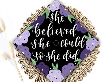 May include: Black graduation cap decorated with purple and green flowers. The cap has the text "She believed she could so she did" written in white.