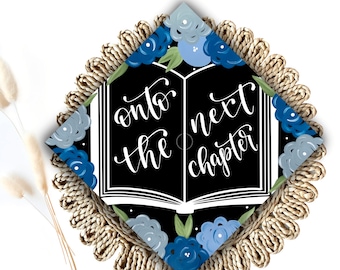 May include: A black graduation cap with a white open book design and the text "onto the next chapter" in white cursive. The book is surrounded by blue and gray flowers on a black background. The cap is decorated with a brown tassel.