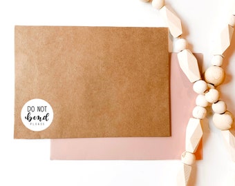 May include: A brown envelope with a white sticker that says "Do Not Bend Please" on it. The envelope is on top of a pink envelope. A white wooden bead garland is in the upper right corner of the image.