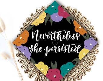 May include: Black graduation cap with a floral design in shades of yellow, orange, red, purple, teal and green. The text "Nevertheless she persisted" is written in white on the cap.