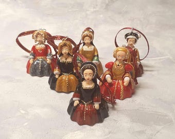 Puede incluir: Un conjunto de seis adornos de campana de cerámica con forma de figuras históricas con vestimenta estilo Tudor. Cada figura lleva un vestido detallado en colores rojo, negro, dorado y marrón, con tocados a juego y una cinta roja para colgar.
