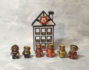 Puede incluir: Una casa decorativa con techo negro y diseño floral rojo y blanco, acompañada de seis pequeñas figuras de osos detalladas. Los osos visten varios atuendos, mostrando una gama de colores y estilos.