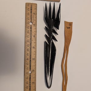 Puede incluir: Un tenedor de madera negra tallada con un diseño dentado, de aproximadamente 18 cm de largo, junto a un pequeño trozo de cuero marrón claro etiquetado "#3". Una regla de madera está en el fondo.