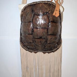 May include: A brown turtle shell handbag with a cream-colored fringe and strap. The bag features a dark brown, textured shell design with visible seams. A small tag with the number "# 4" is attached to the top.