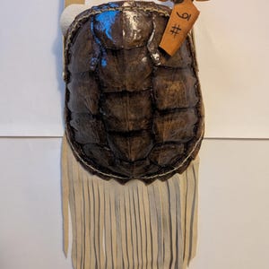 May include: A small, handcrafted bag featuring a turtle shell design. The bag is primarily brown with a textured surface, complemented by a cream-colored fringe along the bottom. A small tag with Japanese text is attached.