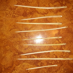 May include: Ten bone needles of varying lengths, arranged vertically on a brown surface.