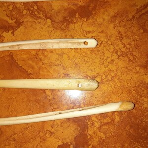 May include: Three wooden knitting needles with a hole at the end of each needle. The needles are on a brown surface.