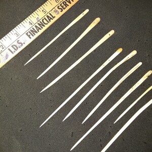 May include: Ten bone sewing needles with rounded tips. The needles are various lengths and are arranged in a diagonal line on a dark gray surface.