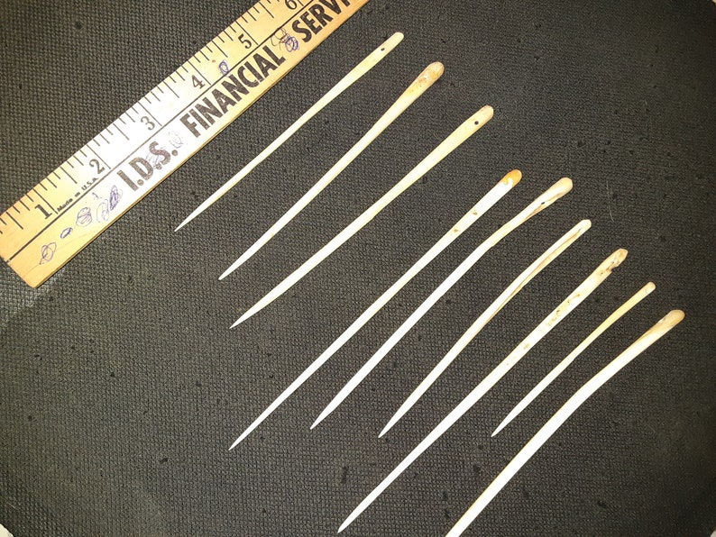 May include: Ten bone sewing needles with rounded tips. The needles are various lengths and are arranged in a diagonal line on a dark gray surface.