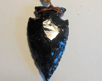 Puede incluir: Un colgante de punta de flecha de obsidiana negra en un cordón de cuero marrón. La punta de flecha está envuelta con alambre de cobre.
