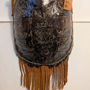 May include: A handcrafted, dark brown turtle shell purse with tan leather accents. The purse features a fringed bottom and a leather strap. The tag on the side reads "#7". The shell has a unique, textured pattern.
