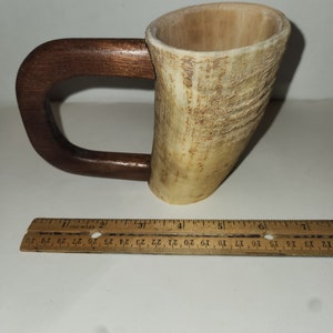 Puede incluir: Una taza de cuerno para beber con un asa de madera, hecha de materiales naturales. El cuerno es de color marrón claro con una textura natural y el asa es de madera marrón oscuro.