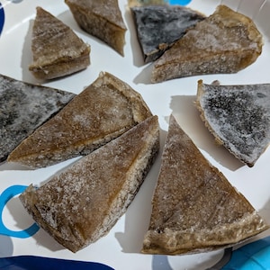 May include: Overhead shot of several triangular food portions, possibly pie or quiche slices, on a white paper plate. The food has a light brown crust and a darker filling, with a dusting of frost or ice crystals. The plate has blue and white patterns.