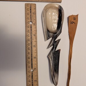 Op de afbeelding: Een handgemaakte lepel met een witte ovale kom en een zwart-wit handvat. Een houten liniaal ligt naast de lepel, met metingen in centimeters. Een klein, dun bruin stuk hout staat ook op de afbeelding, met het nummer 6 erop.
