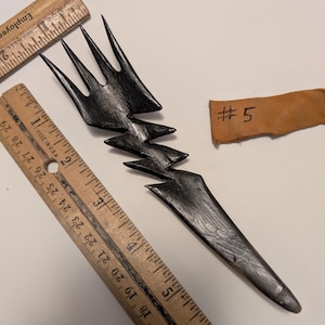 Puede incluir: Un objeto en forma de rayo estilizado de color gris oscuro con una parte superior en forma de tenedor. El objeto está junto a dos reglas de madera y un pequeño trozo de cuero con el número "#5" escrito. Las reglas están marcadas en pulgadas.