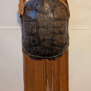 Puede incluir: Un bolso de hombro único, hecho a mano, con un cuerpo de caparazón de tortuga y flecos de cuero marrón. El bolso tiene una correa de cuero y una etiqueta. El caparazón es marrón oscuro y texturizado, y los flecos son de color marrón claro.