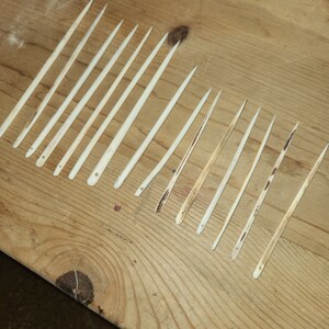 May include: A collection of 18 natural bone knitting needles, arranged in rows on a wooden surface. The needles are various lengths and have a natural, light brown color.
