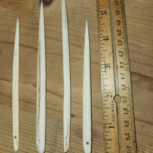 May include: Four white bone awls of varying lengths, measured against a wooden ruler marked in inches. The awls are sharpened at one end and have a small hole at the other end.