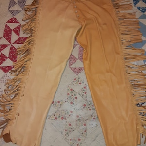 May include: A pair of light brown leather trousers with fringe detailing on the sides. The trousers have a button and zip fastening at the front.