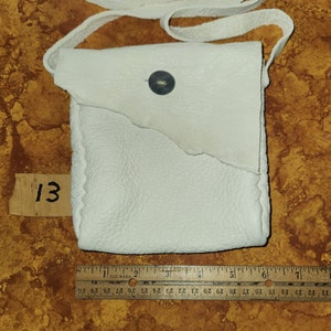 Puede incluir: Un bolso bandolera de cuero blanco con un cierre de botón único. El bolso tiene un diseño sencillo y está hecho de cuero suave y flexible.