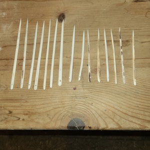 May include: A collection of 14 thin, white, bone needles laid out on a wooden surface.
