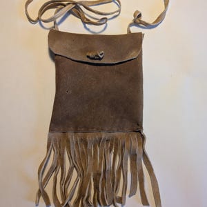 Puede incluir: Una bolsa de cuero marrón de gamuza con flecos y una correa de cuero. La bolsa tiene una solapa con cierre de botón.