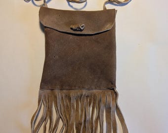 May include: A brown suede leather pouch with a fringe trim and a leather strap. The pouch has a flap with a button closure.