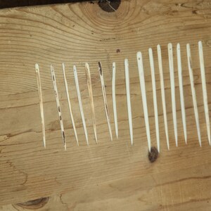 May include: A collection of 14 thin, wooden needles, laid out in a row on a wooden surface. The needles are all the same color, a light beige, and are slightly different lengths.