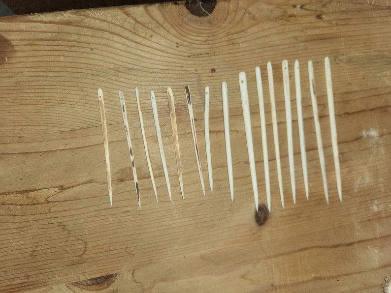 May include: A collection of 14 thin, wooden needles, laid out in a row on a wooden surface. The needles are all the same color, a light beige, and are slightly different lengths.