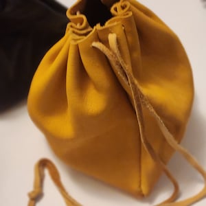 Puede incluir: Una pequeña bolsa de cuero amarilla con cierre de cordón. La bolsa está hecha de cuero suave y flexible y tiene un aspecto rústico y artesanal.