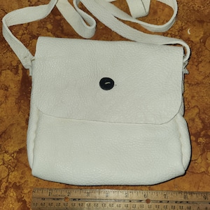 Puede incluir: Un bolso bandolera de cuero blanco con un cierre de botón único. El bolso tiene una correa larga y está hecho de cuero texturizado.